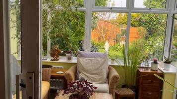 A cozy room with a wicker chair, a small table with a vase of flowers, and a view of a garden through large windows.