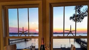 A dining table with a bottle of champagne, two chairs, and a view of the sunset over the water through large windows. -  WATERFRONT Property on KDH Bike Path with water access, bikes, kayak, and canoe (Kill Devil Hills)