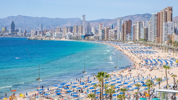 Un paisaje urbano costero con una playa bordeada de sombrillas azules y palmeras.