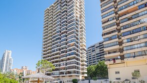Un edificio residencial alto con varios balcones, un jardín en la azotea y una zona de piscina con sombrillas.