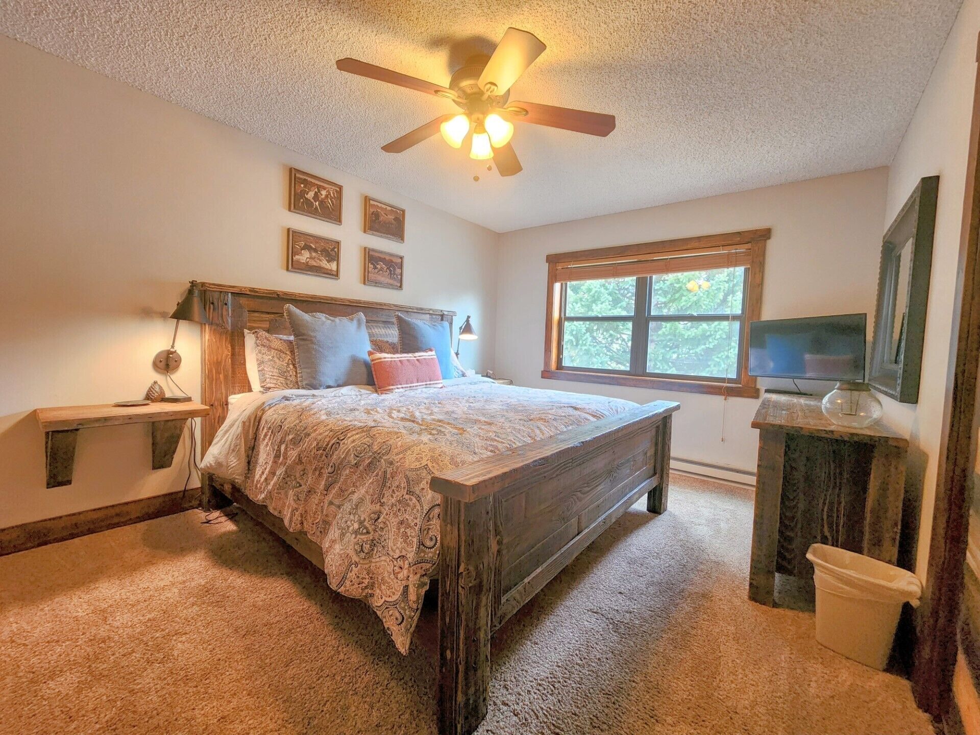 Une chambre avec un lit en bois, un ventilateur de plafond, une fenêtre et un téléviseur posé sur un meuble en bois.