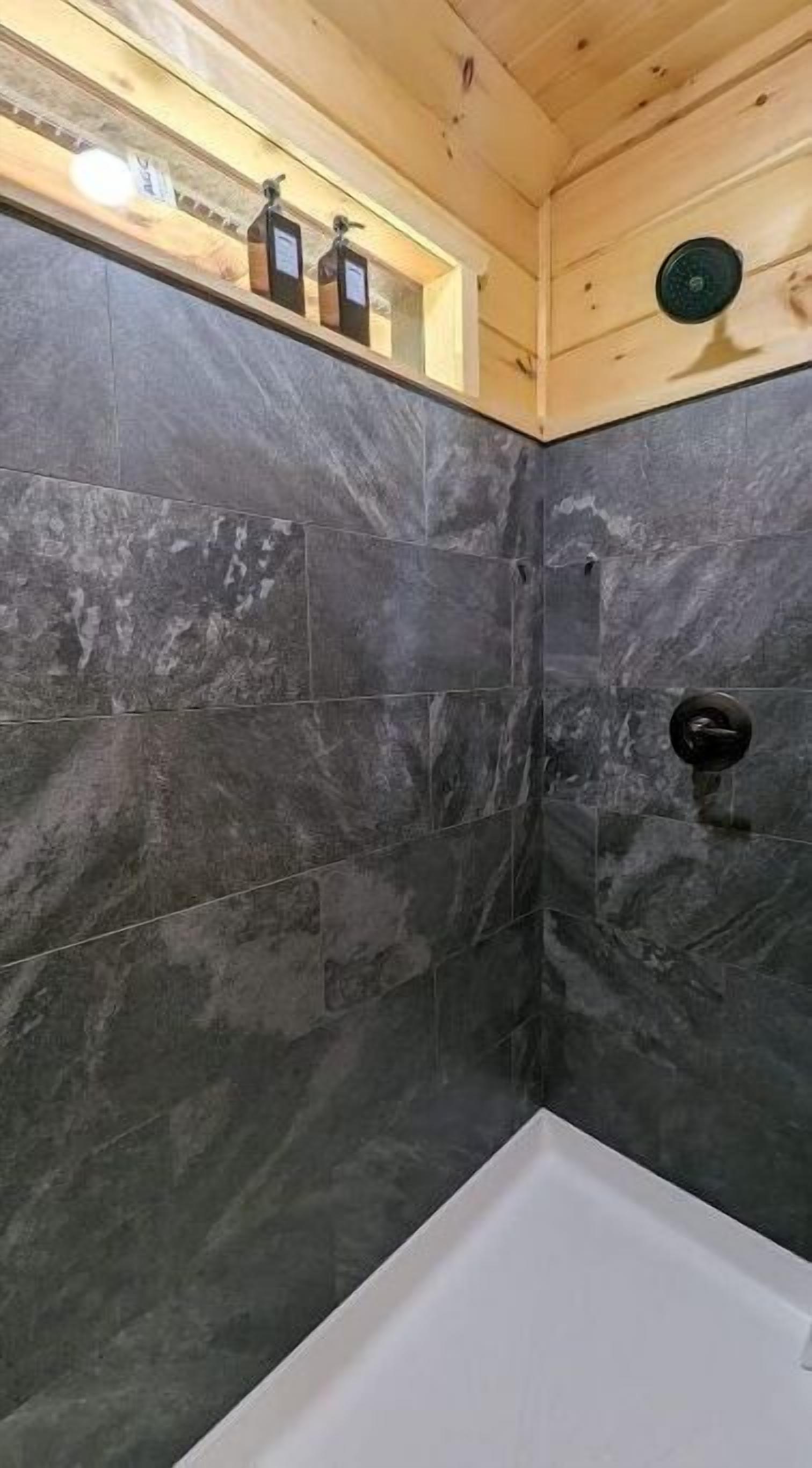 Un baño con baldosas de mármol oscuro, una bañera blanca y techo de madera.