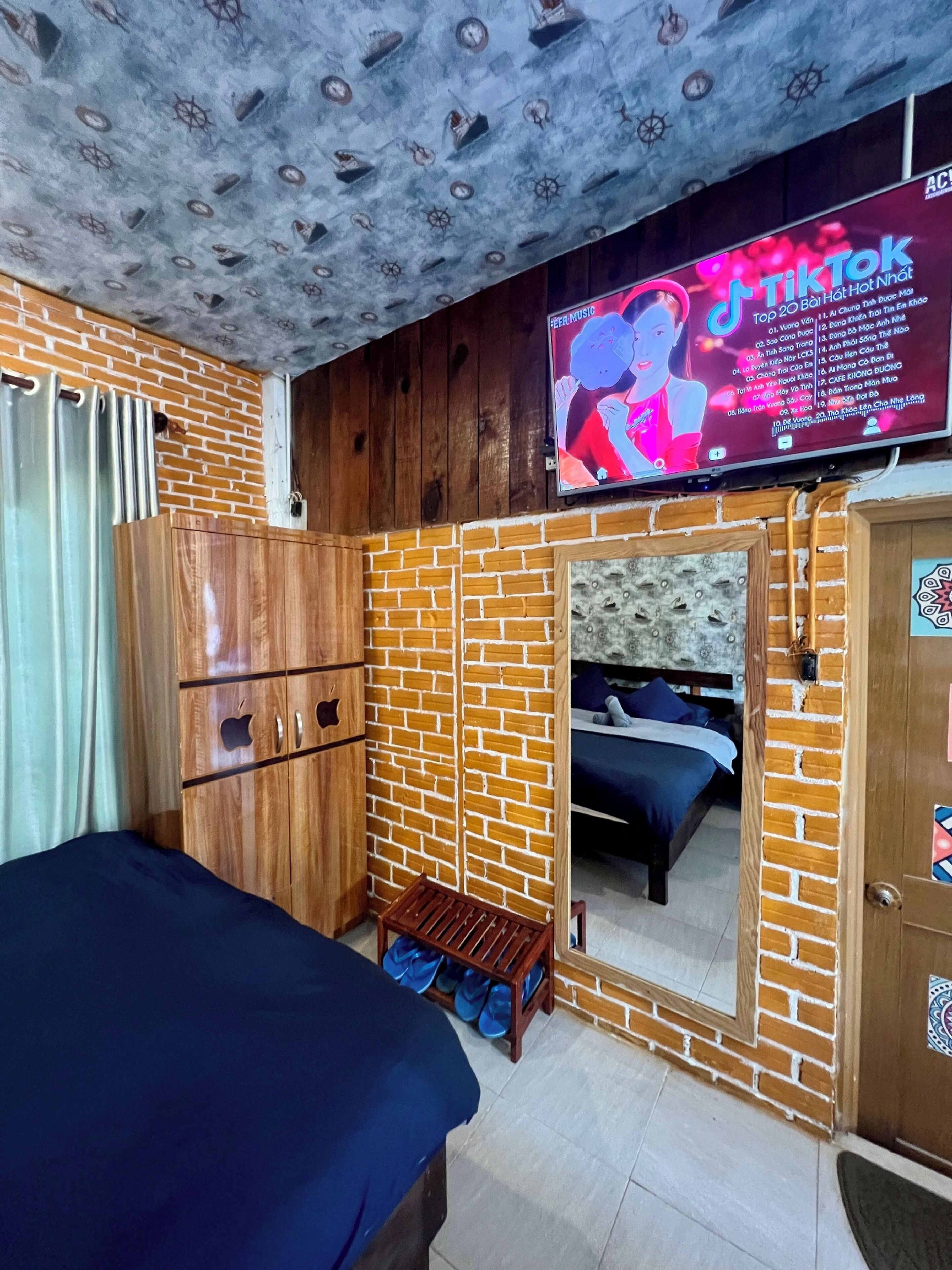Une pièce avec un mur décoré d’un motif en briques, une armoire en bois, un lit, un téléviseur fixé au mur et un petit tabouret en bois.