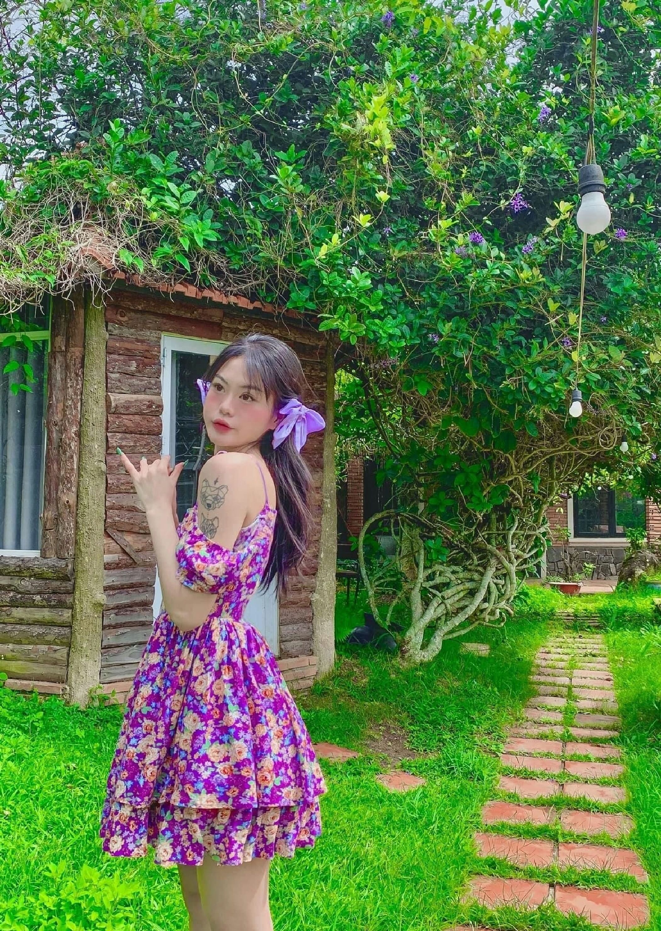 A person in a floral dress standing in front of a rustic cabin with a garden path.