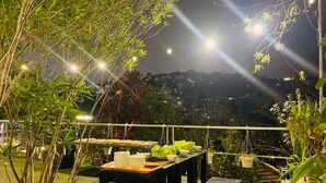 Un espacio de comedor al aire libre con mesas y sillas, rodeado de follaje e iluminado con luces colgantes.