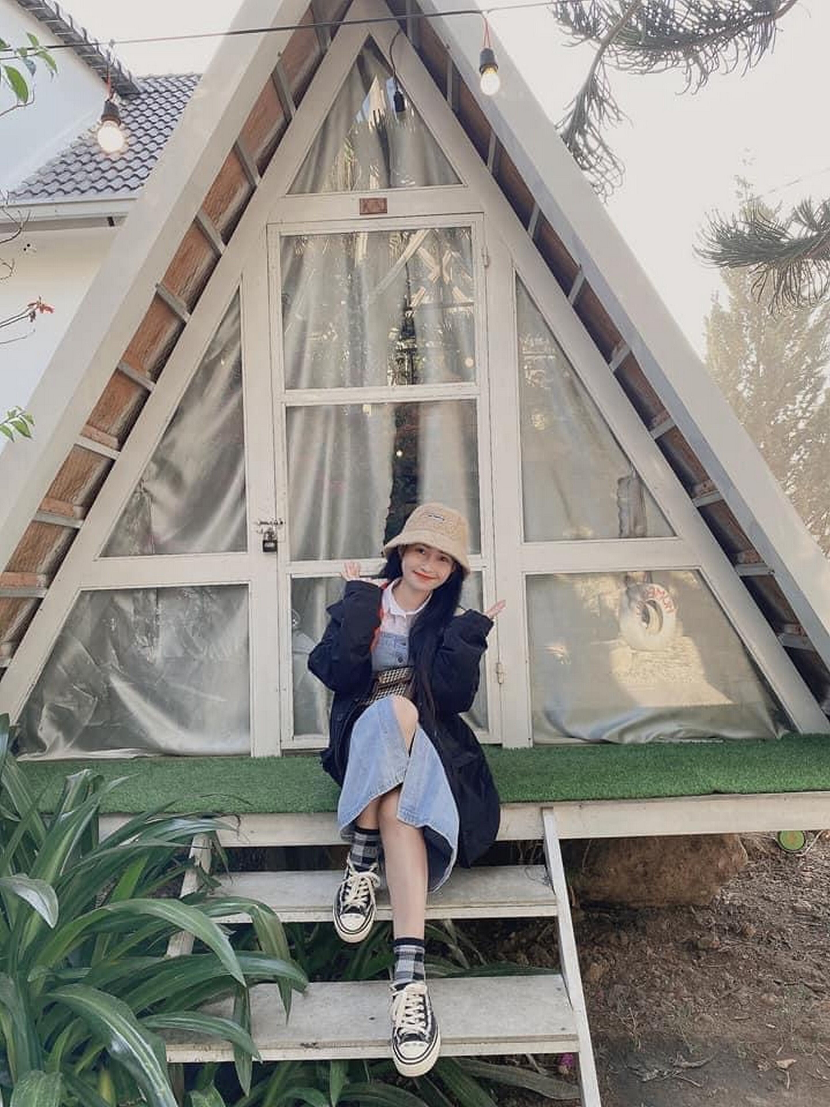 A person sitting on a wooden bench outside a building with a triangular roof and a large window.