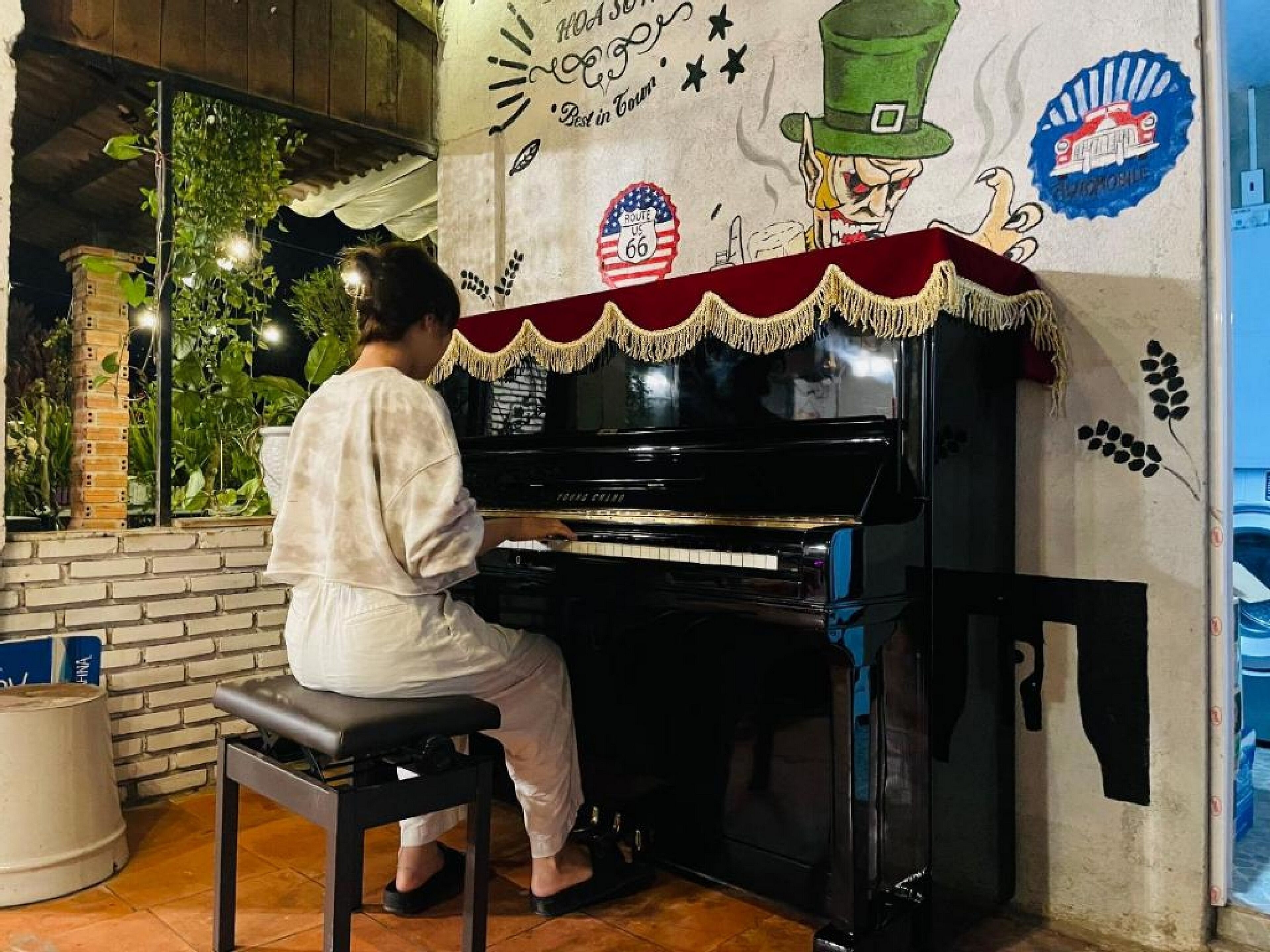 A person playing a piano in a room with a mural, brick wall, and potted plants.
