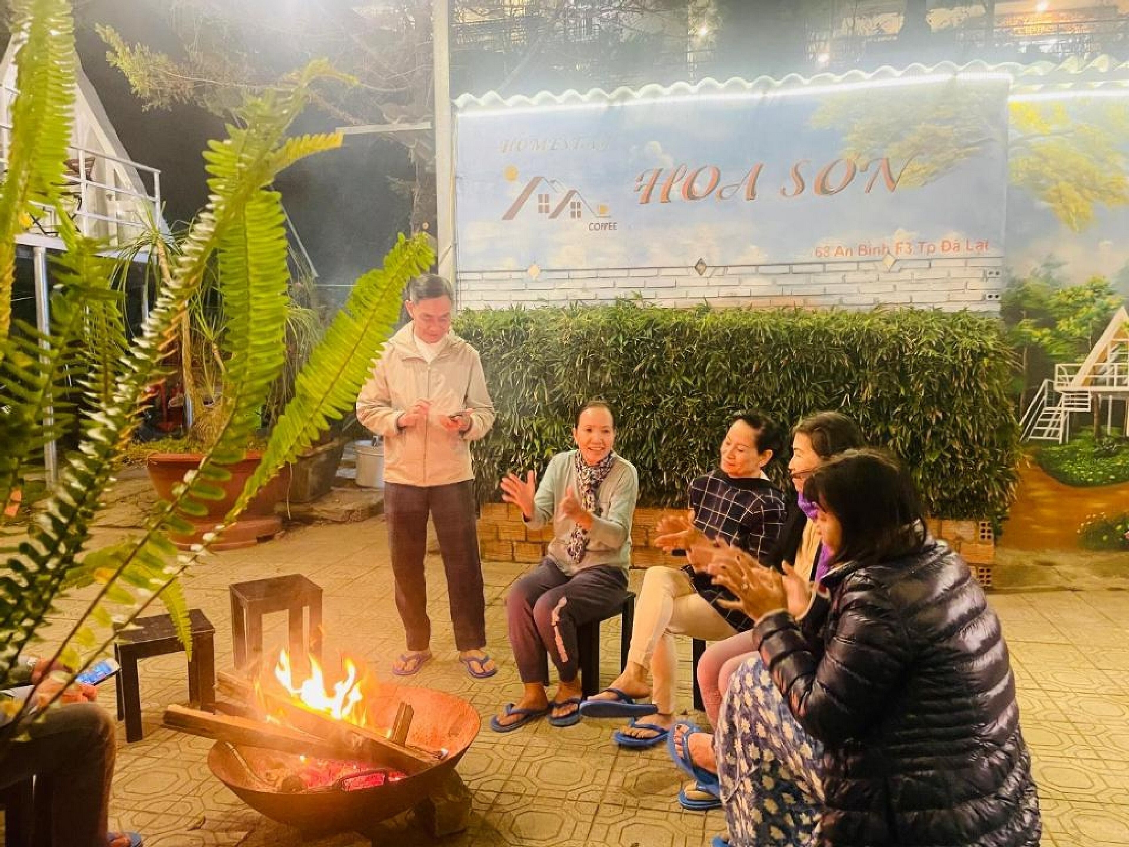 A group of people sitting around a fire pit in an outdoor setting with lush greenery and a large banner in the background.