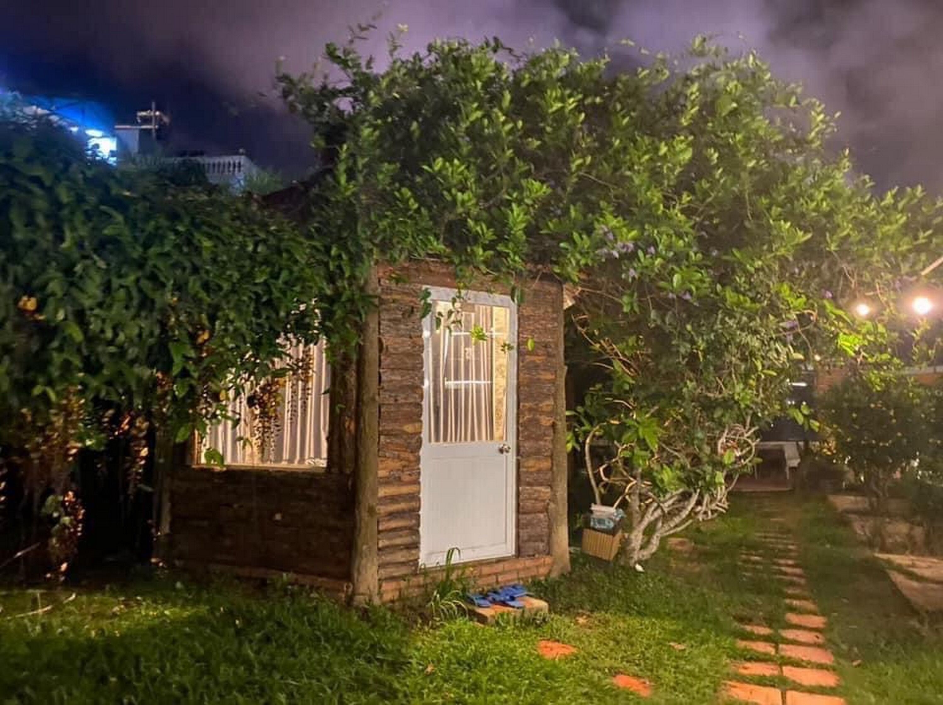 A small, rustic wooden structure with a window and a door, surrounded by lush greenery and a paved path.