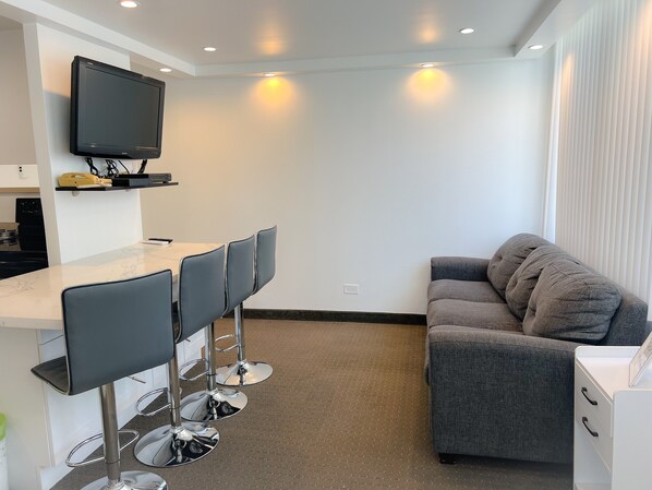 A compact living space with a kitchenette, a dining area with bar stools, a grey sofa, and a flat-screen TV mounted on the wall.