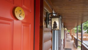 Una puerta roja con una placa de números, un farol con silueta de árbol y un porche de madera con barandilla.