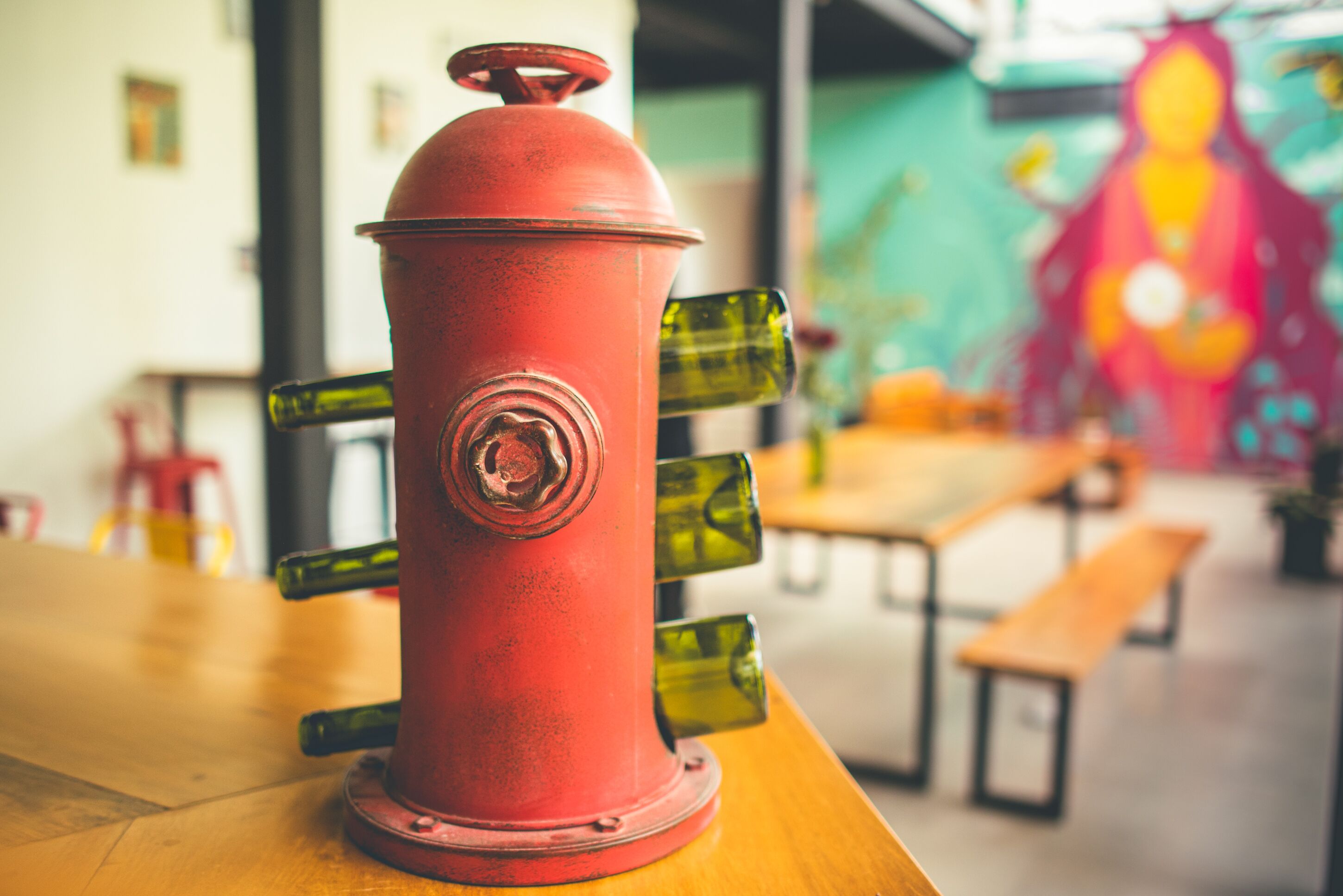 Um hidrante vermelho com várias bocas de vidro verde sobre uma mesa de madeira num espaço com bancos e um mural na parede.