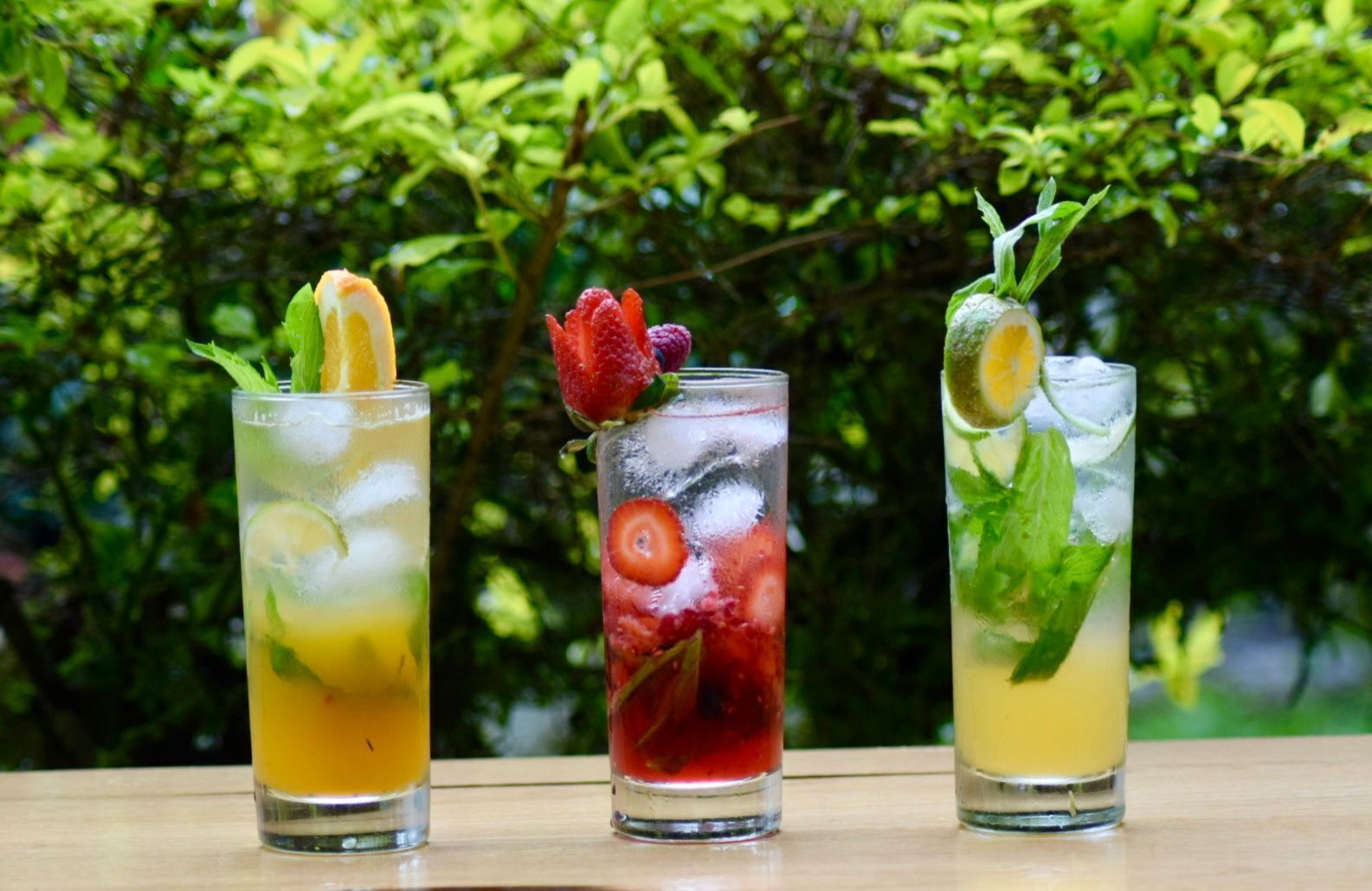 Three glasses of iced drinks with fruit garnishes on a wooden table against a green leafy background.