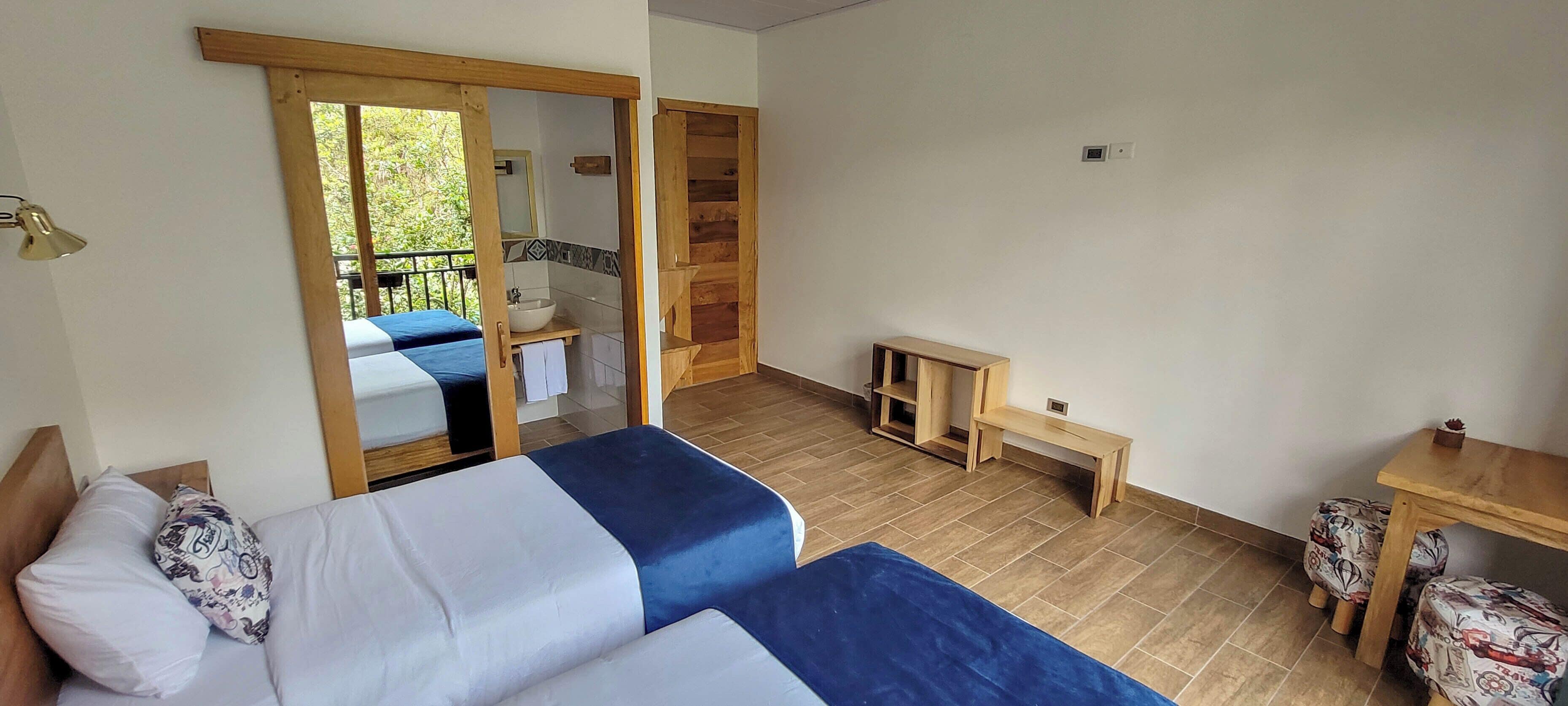 A room with two beds, a wooden bench, a small table, and a bathroom visible through an open door.