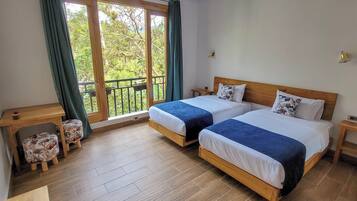 A bedroom with two beds, a wooden nightstand, a small table, and a view of greenery through a window.