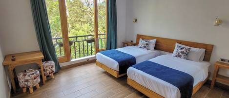 A bedroom with two beds, a wooden nightstand, a small table, and a view of greenery through a window.