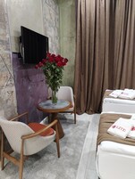 Una habitación de hotel con una cama, una mesita redonda con un jarrón de rosas rojas, dos sillas, un televisor montado en la pared y una ventana con cortinas.