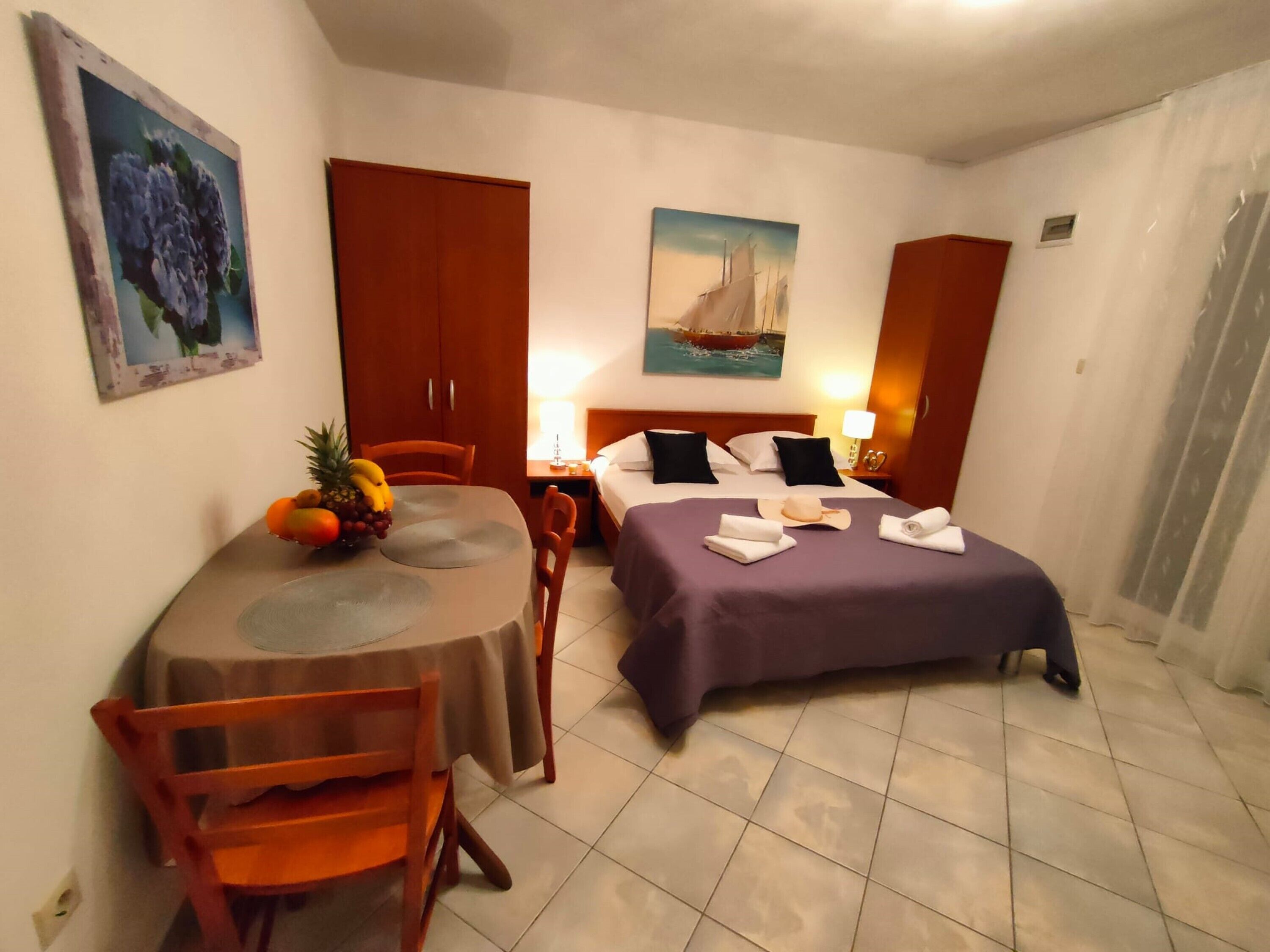 A hotel room with a bed, a dining table with chairs, a fruit bowl, and artwork on the walls.