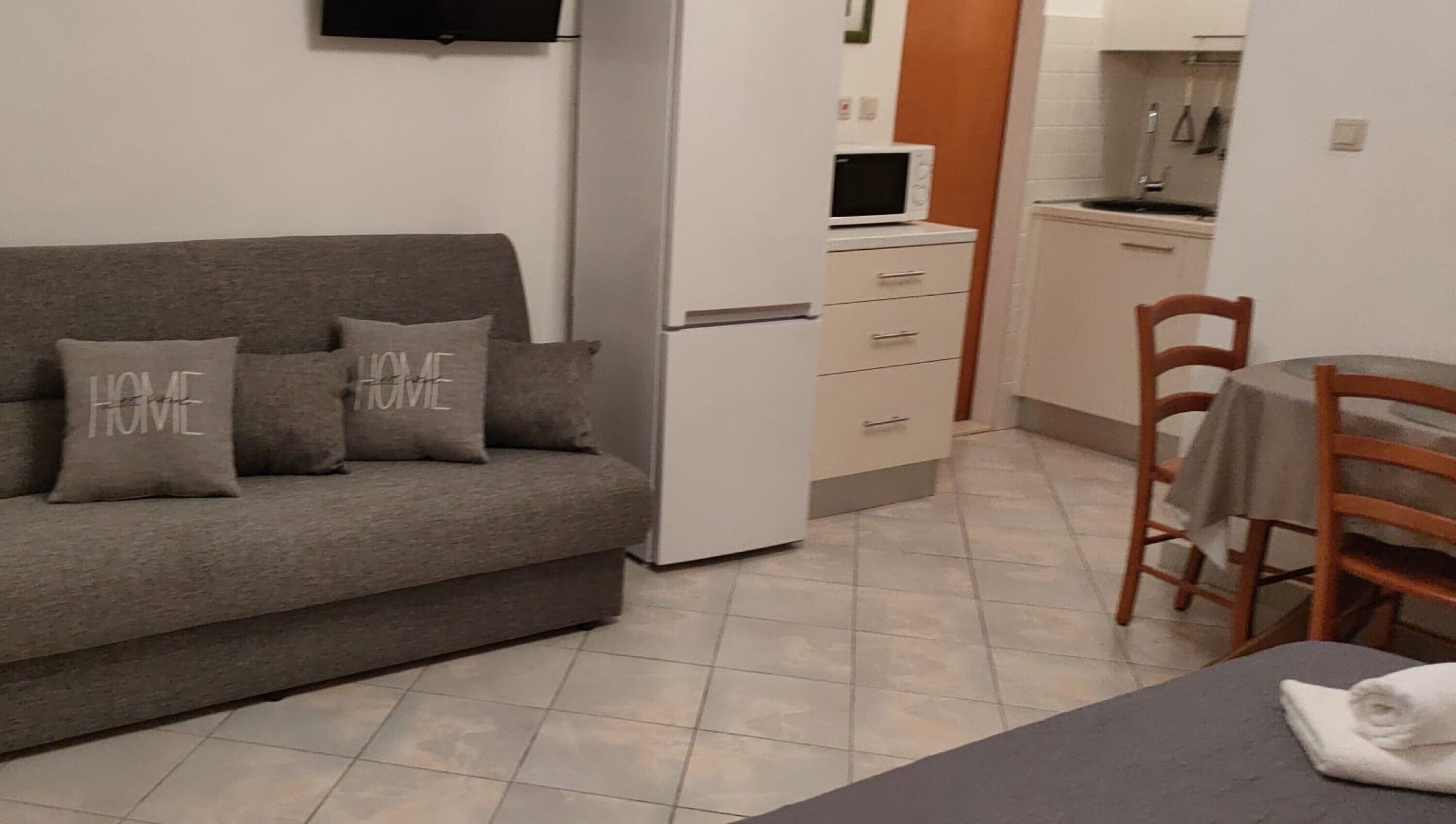 Un espace de vie compact comprenant un canapé gris, deux coussins ornés du mot « HOME », un petit coin cuisine avec un four à micro-ondes et une table à manger avec une chaise.