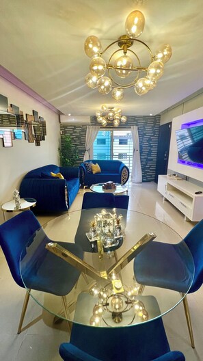 A modern living room with a glass coffee table, blue sofas, a TV, and a chandelier.