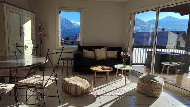 A spacious living area with a large window offering a mountain view, a black sofa, a dining table with chairs, and a small round table with a vase. - Résidence Calme à Proximité Immédiate D'un Sentier de Randonnée (Embrun)