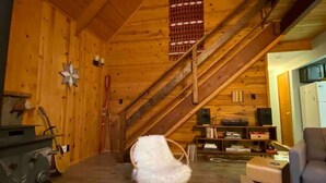 A cozy wooden interior with a stone fireplace, a white chair, a wooden table, and a staircase leading to an upper level. - Cozy Mt. Baker Cabin, Wi-Fi, Sleeps 6-8, Pets OK (Deming)