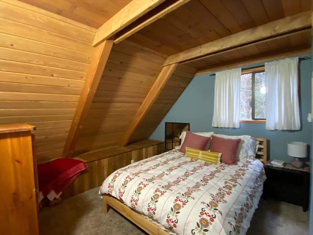 A bedroom with a sloped ceiling, a bed with floral bedding, a nightstand with a lamp, and a window with curtains.