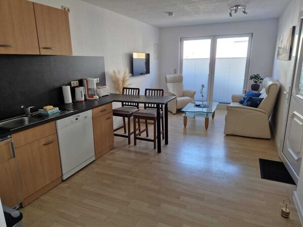 Una cocina compacta con electrodomésticos modernos, un comedor con una mesa de centro de vidrio y una sala de estar con sofá y planta.