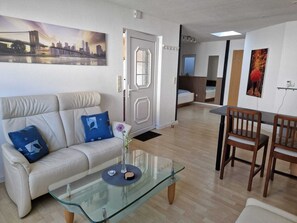 Una sala moderna con una mesa de centro de vidrio, un sofá color crema con cojines azules, una zona de comedor con sillas de madera y un cuadro de un paisaje urbano en la pared.