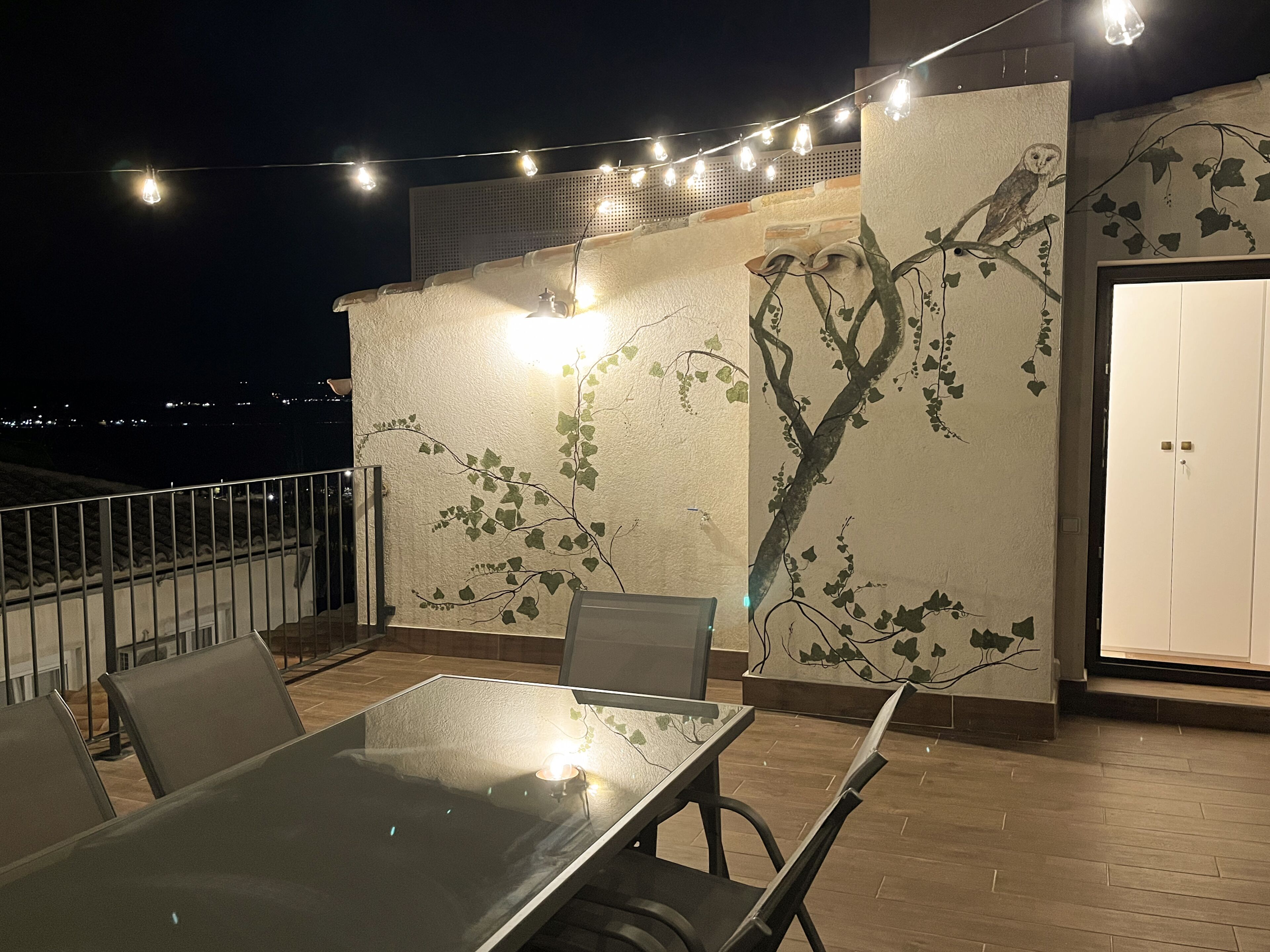 An outdoor dining area with a glass table and chairs, string lights, and a wall mural of a tree with vines.