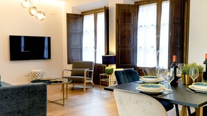 Una sala de estar moderna con un televisor de pantalla plana, asientos cómodos, una mesa de comedor para cuatro personas y ventanas amplias con cortinas.