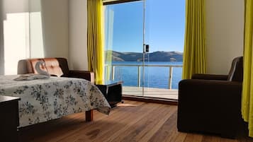 A room with a bed, a desk, and two chairs, with a view of a body of water and hills.