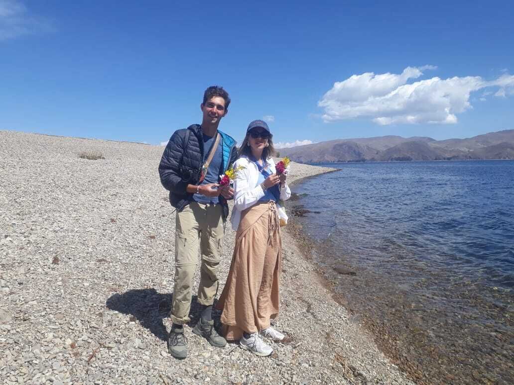 Two people standing on a pebbled shore by a lake with mountains in the background.