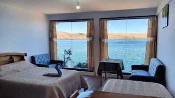 A hotel room with a bed, a chair, a small table, and a sofa, all facing a large window with a view of a body of water and hills.