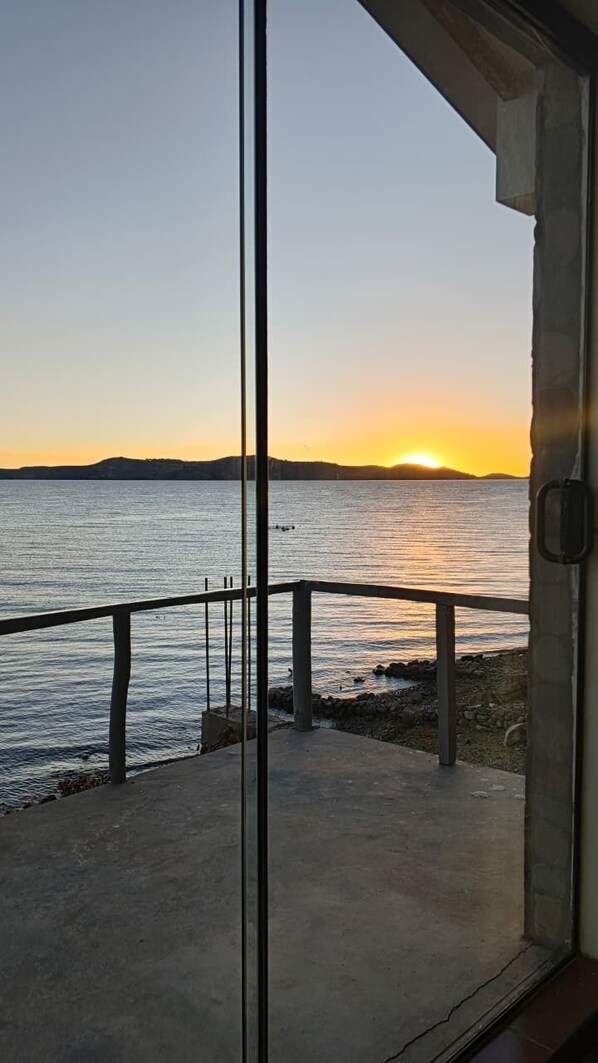 Vista de un atardecer sobre el agua desde un balcón con piso de concreto y baranda de metal.