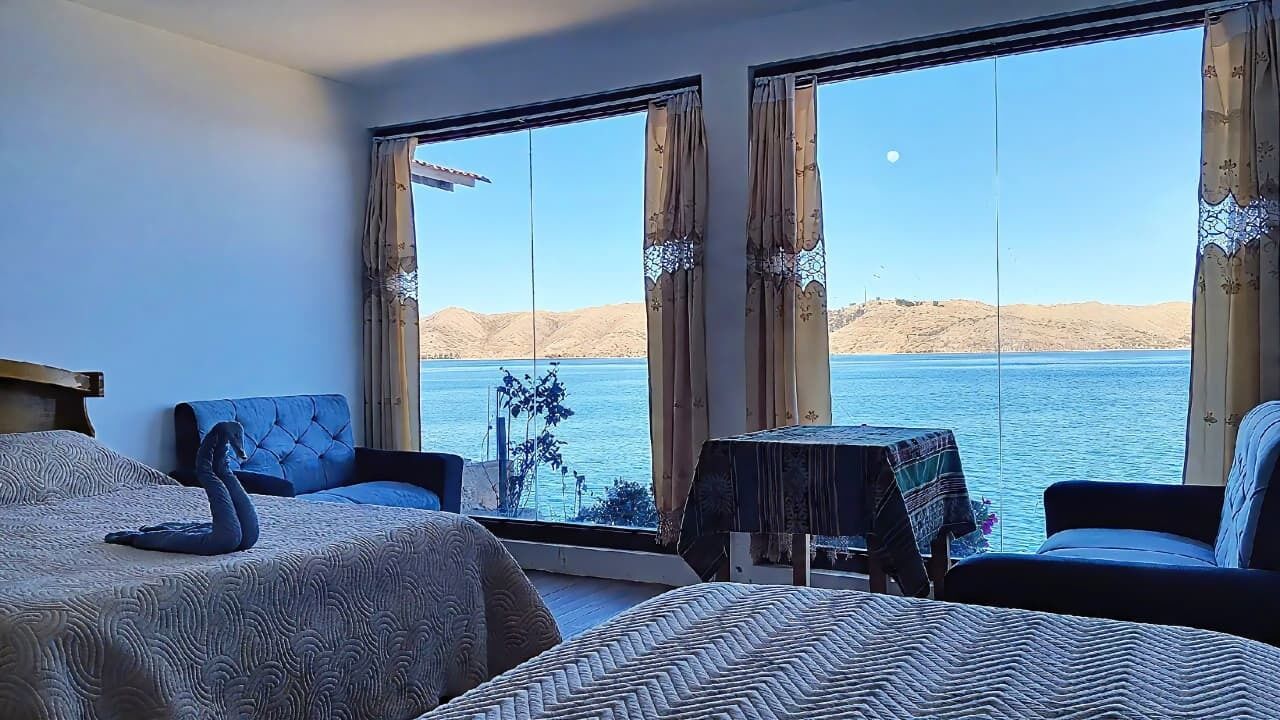 A room with two beds, a table, and a view of a body of water with hills in the distance.