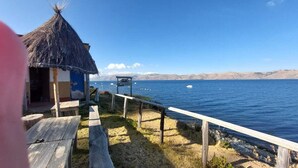 A wooden deck with a thatched-roof structure, a sign reading "Hostal," and a view of a calm lake with boats. - LUNA DEL TITIKAKA LODGE (Moon Island)