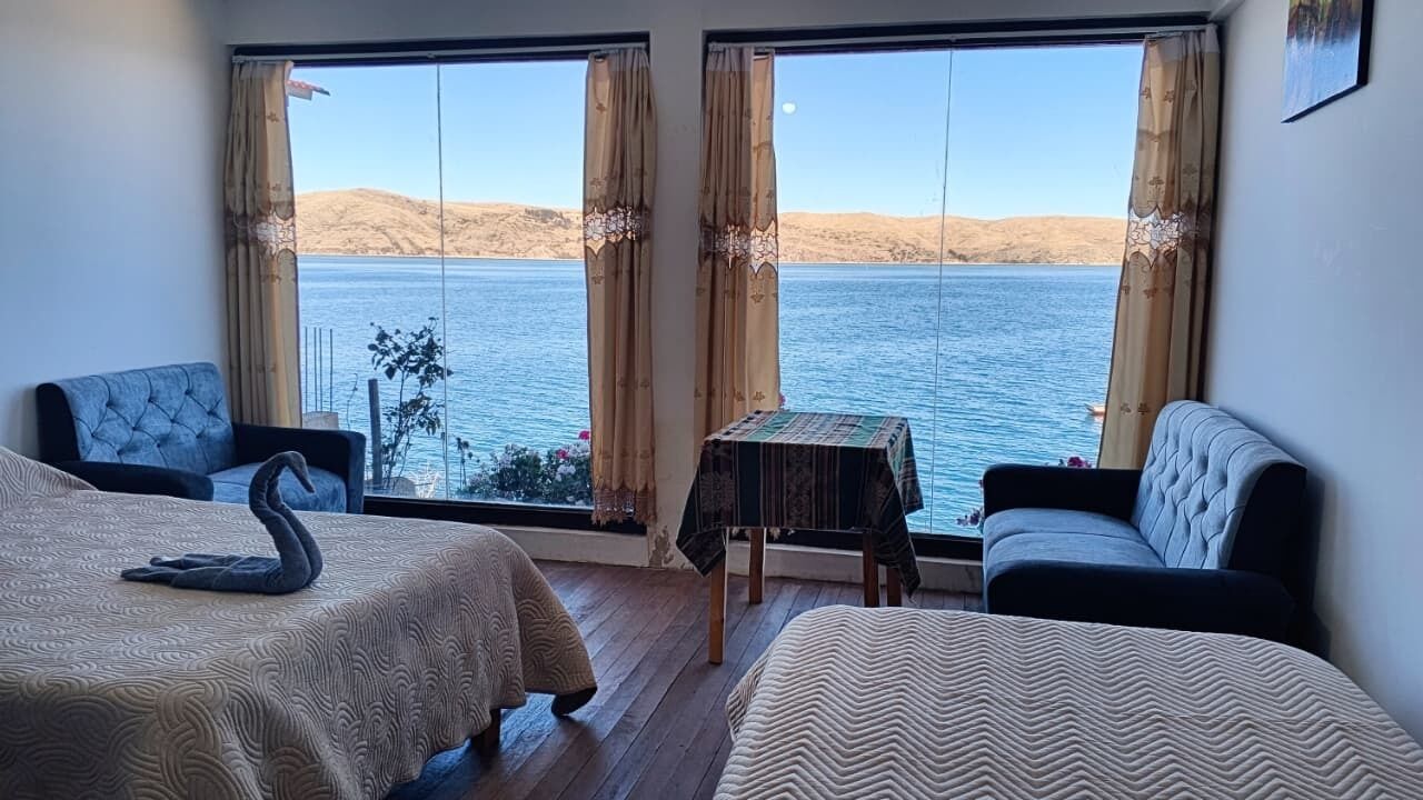 Habitación de hotel con dos camas, un sofá y una mesa, todos mirando a un amplio ventanal con vistas a un lago y colinas.