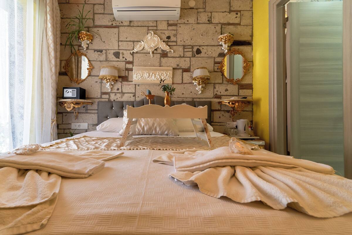 A hotel room with a brick wall, a bed with a white headboard, a small shelf with a radio, and a mirror.