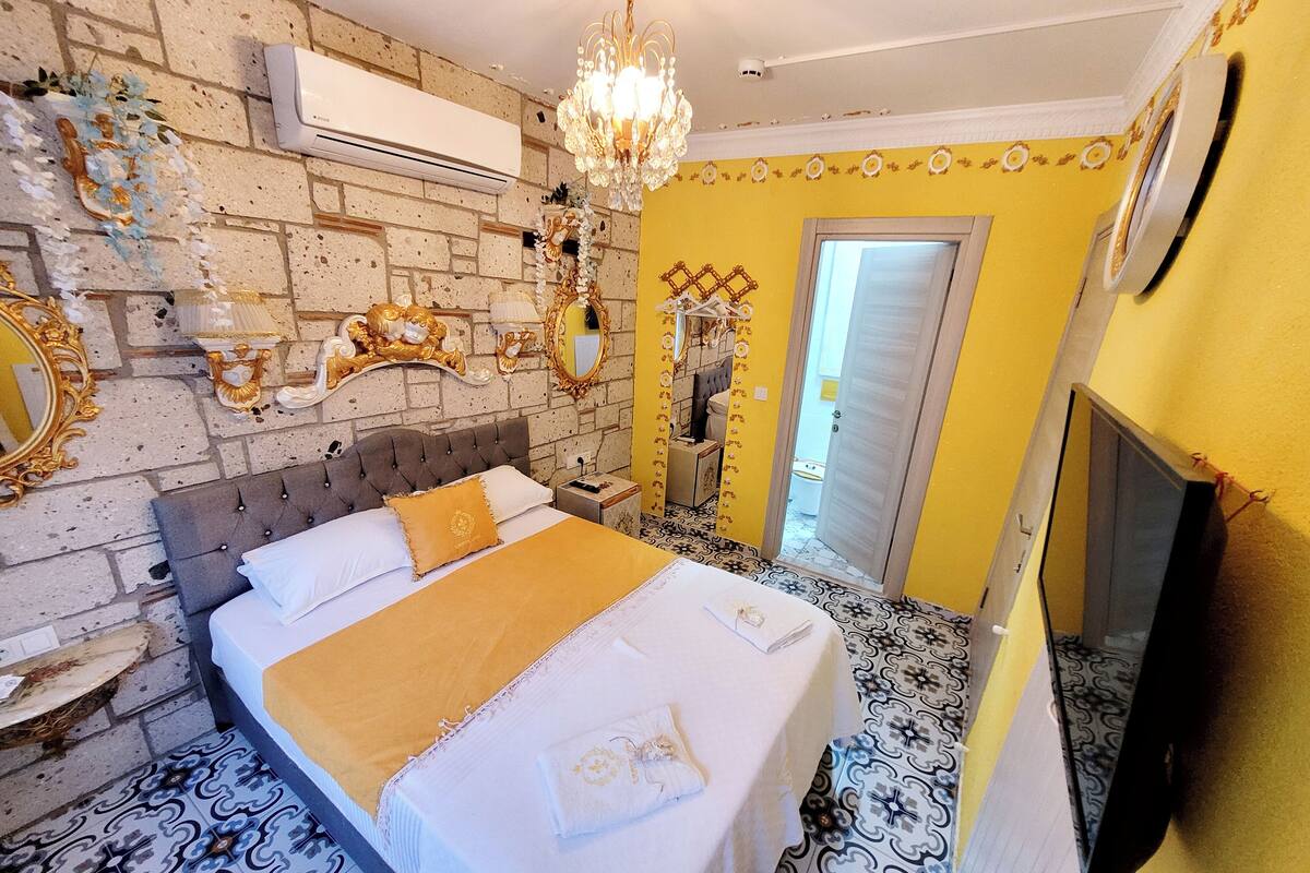 A bedroom with a brick wall, a chandelier, a bed with a gold throw, and a TV on the wall.