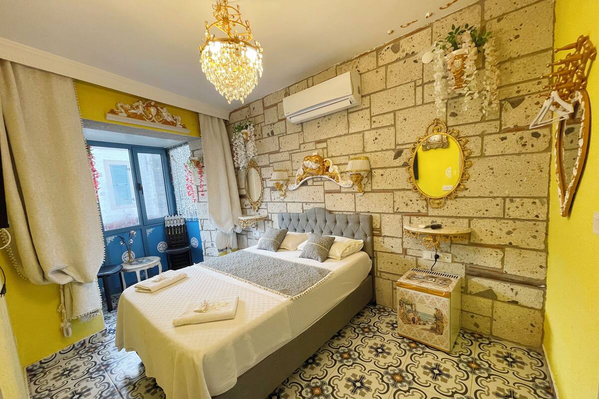 A bedroom with a large bed, a chandelier, a mirror, and a brick-patterned wall.