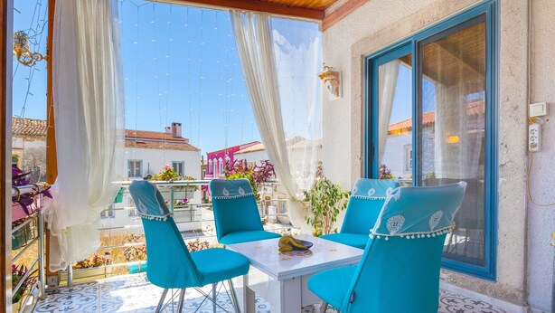 A sunny terrace with turquoise chairs and a table, overlooking a street with buildings and flowers. - Lamur Otel (Cesme)