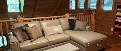 A cozy wooden cabin interior with a sectional sofa and ottoman, wooden floors, and a wooden railing.