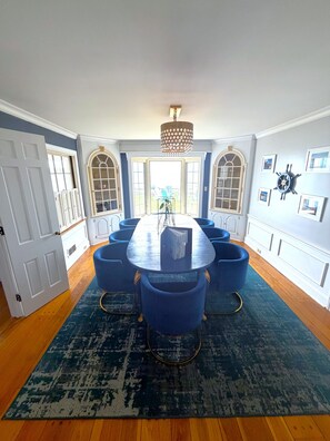 A dining room with a round table, blue chairs, a chandelier, and large windows.