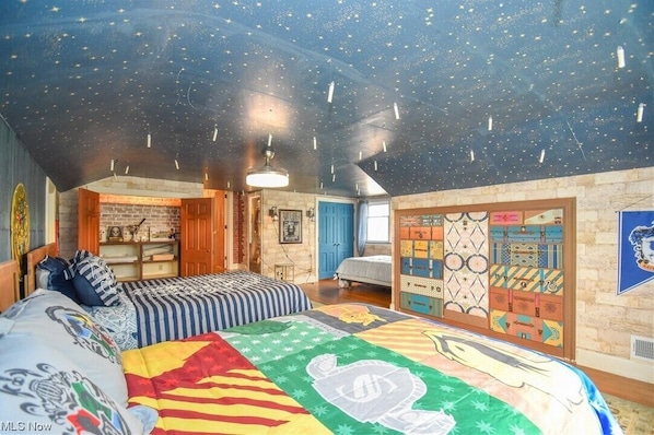 A room with a sloped ceiling, a bed with a colorful quilt, a wall with a suitcase display, and a brick wall.