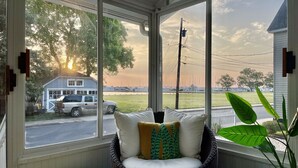 A room with a large window overlooking a street, a wicker chair with cushions, a potted plant, and a view of a house and parked vehicles. - New listing! Riverside Retreat on the Choptank River (Cambridge)