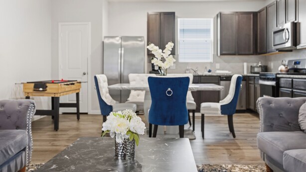 A modern living room with a dining table, sofas, a coffee table with flowers, a kitchen area with cabinets and appliances, and a door. - Lux New Beau;Houston;IAH Airport;Dtown;Woodlands;Spring;NRG World Cup Std;HotTub (Houston)
