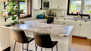 Una cocina moderna con armarios blancos, una isla grande con dos taburetes de bar, una planta colgante y un ventanal con vistas al exterior.