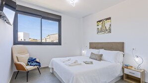 Un dormitorio con cama, una silla, una mesita de noche, una lámpara y una ventana con vista a la ciudad.