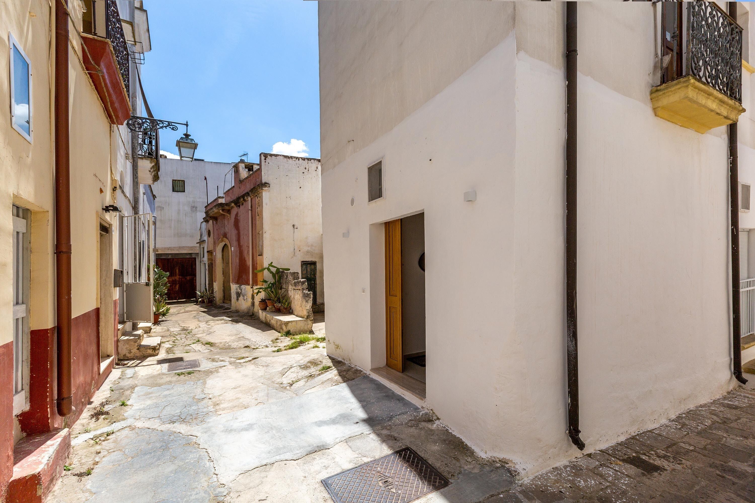 Un estrecho callejón flanqueado por edificios, con una rejilla metálica en el suelo y una puerta de madera a la derecha.